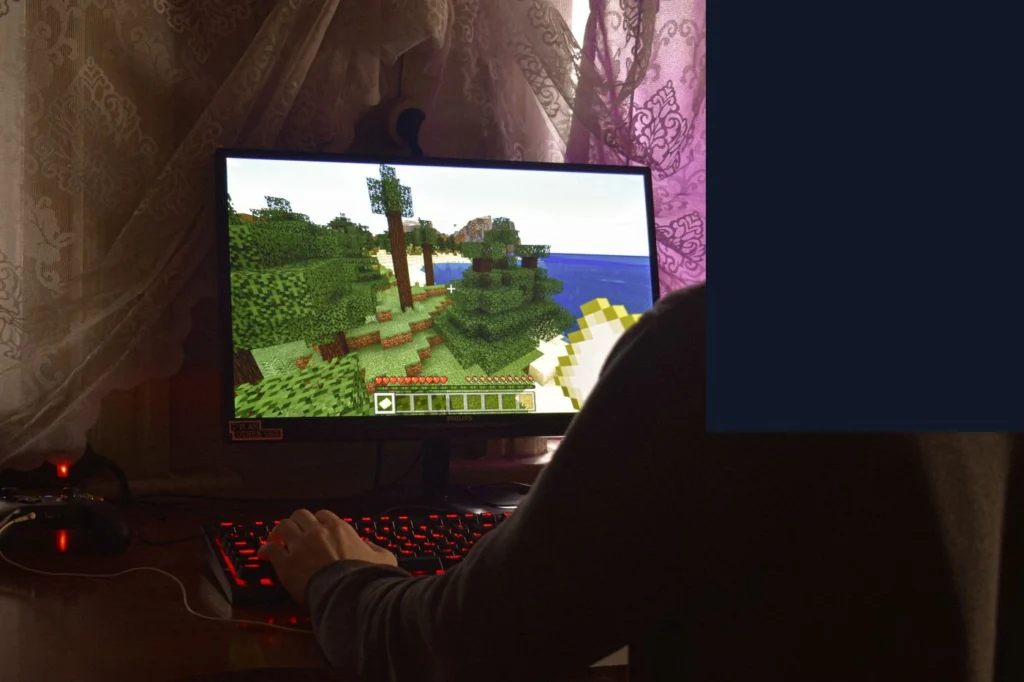 Bilgisayar ekranında Minecraft oyunu oynanıyor; ekranda ağaçlık/orman arazisi ve envanter çubuğu görünüyor, oyuncu klavyede