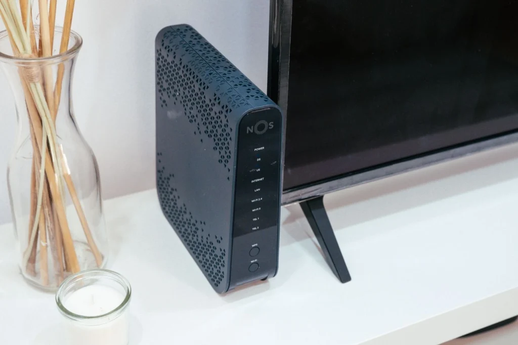 TV ünitesi üzerinde duran NOS marka modem/router, ön panelinde LED ışıkları ve güç/bağlantı göstergeleri görünüyor