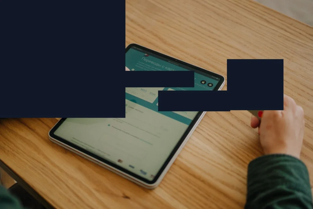 Bir tablet ekranında ödeme formunun açık olduğu, elinin kart bilgisi giriş alanlarını işaret ettiği, ahşap masada kredi kartı bulunduğu görsel