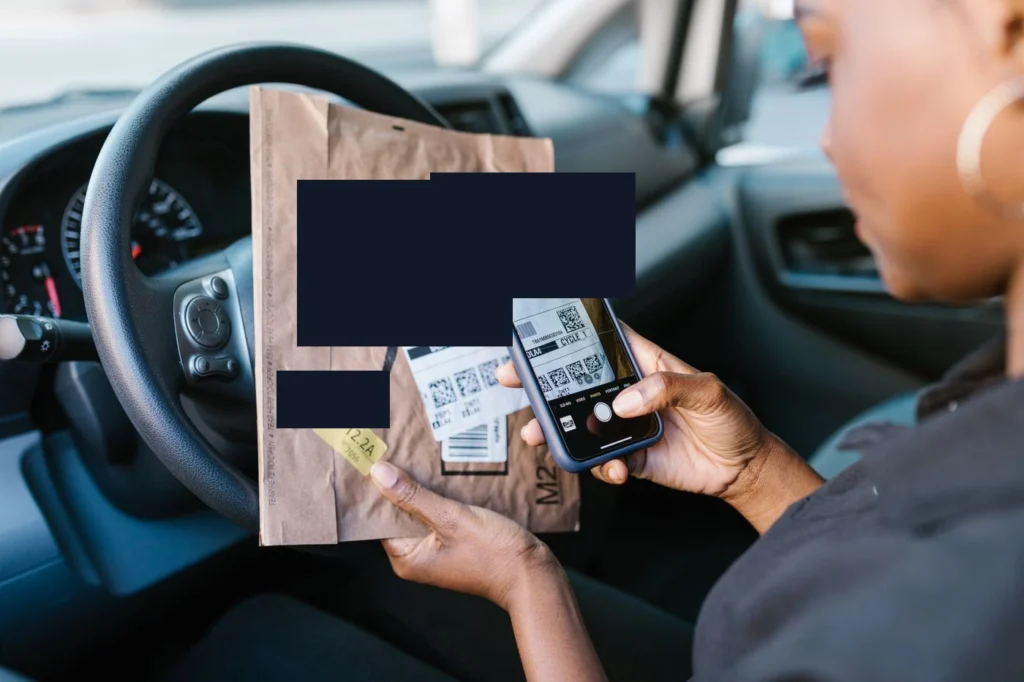 Bir kişi araç içinde kargo paketi ve telefonunu elinde tutarak paketin etiketini kontrol ediyor, kargo takip işlemini yapıyor