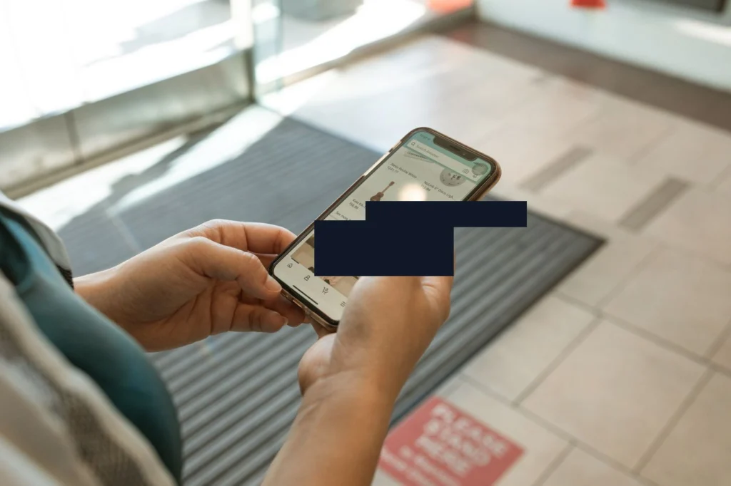 Bir smartphone'un elinde tutulduğu görüntü. Ekranda e-ticaret uygulaması açık, sipariş detayları ve teslimat adresi bilgileri görünüyor. Düz bir yüzey (kachel/zemin) arka planda bulanık şekilde görülü