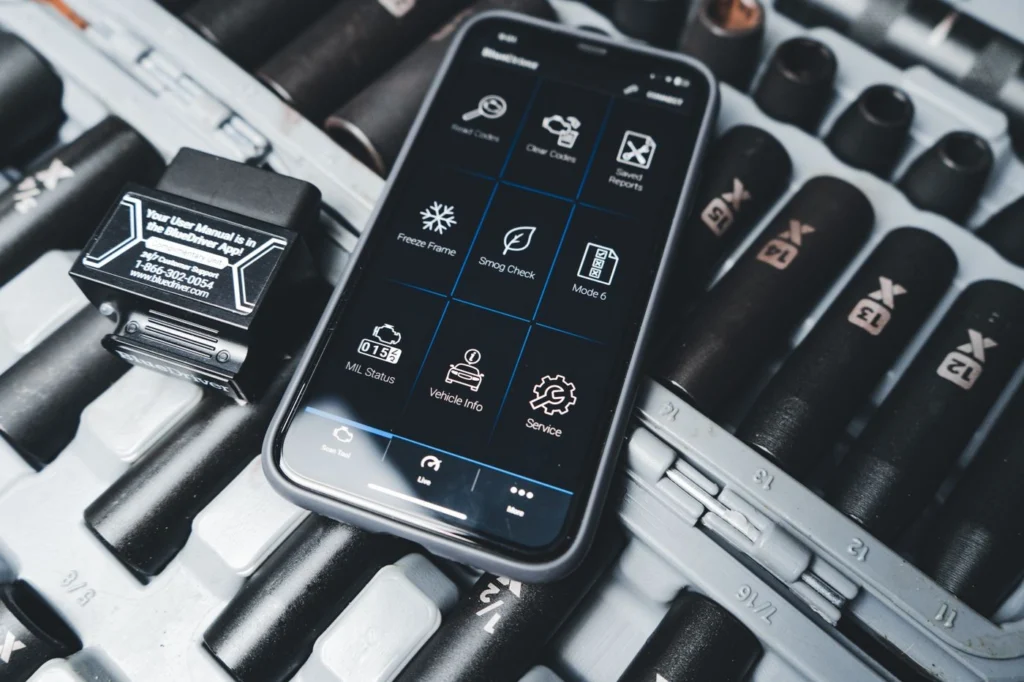 Bir akıllı telefon, bir araç kontrol paneli (OBD uygulaması benzeri) arayüzü gösteriyor. Ekranda arıza teşhisi ile ilgili simgeler ve seçenekler bulunmaktadır.