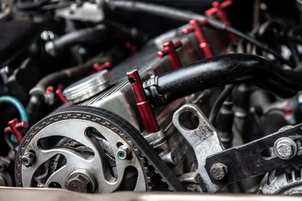 Araç motor bölmesinde kırmızı braketler, siyah hortumlar ve metalik motor parçaları görülmektedir. Direksiyon pompası kayışı bölgesinin detaylı fotoğrafı.